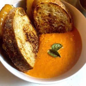 Bowl of homemade creamy tomato soup. Its a rich orange color from the variety of tomatoes used and garnished with fresh basil leaves and cracked black pepper. There is a decadent sourdough grilled cheese dunked into the soup.