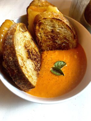 Bowl of homemade creamy tomato soup. Its a rich orange color from the variety of tomatoes used and garnished with fresh basil leaves and cracked black pepper. There is a decadent sourdough grilled cheese dunked into the soup.